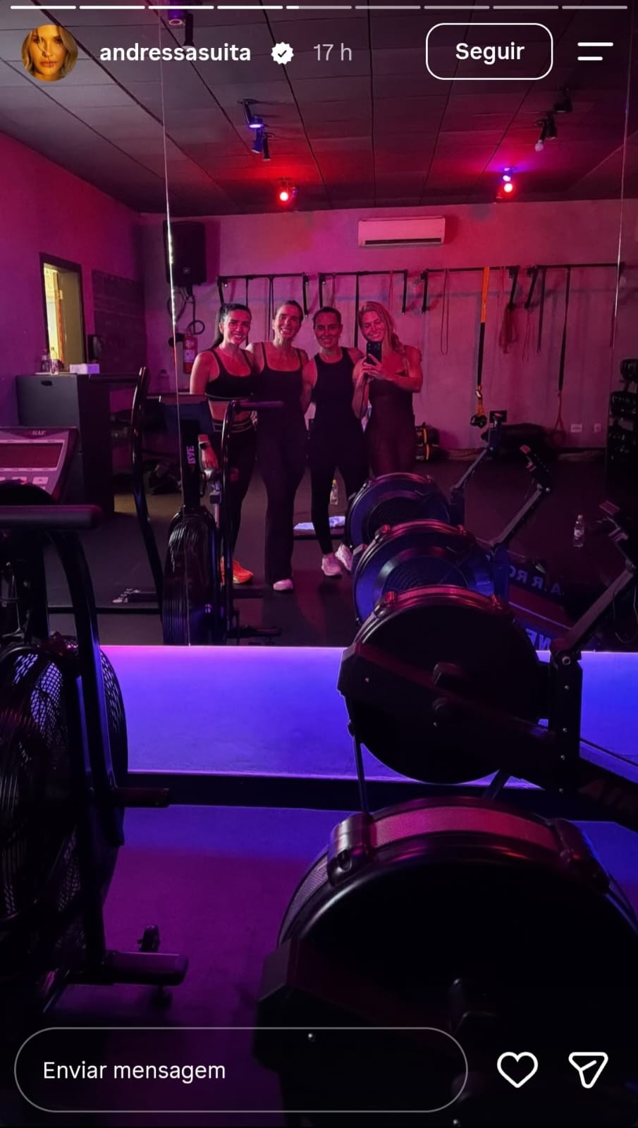 Quatro mulheres sorridentes, vestindo roupas de ginástica escuras, posam para uma selfie em uma academia com iluminação roxa e vermelha. Elas estão refletidas em um espelho, com equipamentos de remo e bicicletas ergométricas em primeiro plano