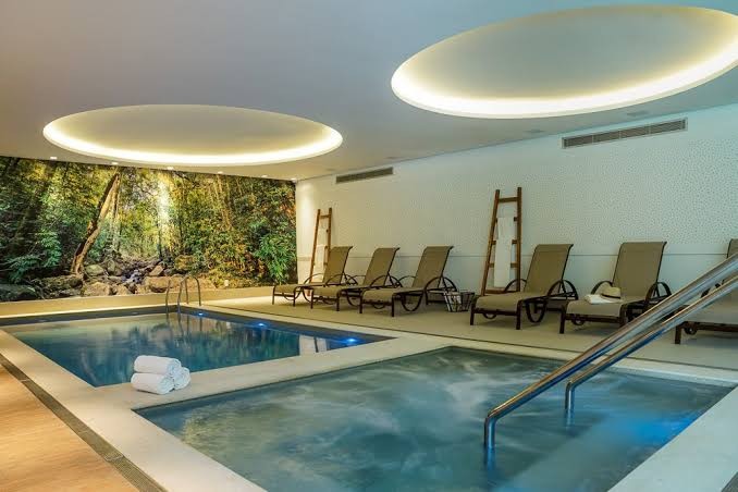 Piscina interna de um spa com água azul clara, ladeada por espreguiçadeiras bege e toalhas brancas. Uma parede tem um mural de floresta tropical, e o teto branco possui duas luminárias ovais com luz amarela suave