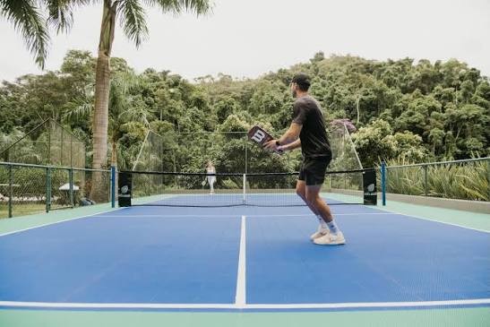 Homem de boné e roupa escura joga pickleball em quadra azul e verde, segurando a raquete. Uma mulher de branco está ao fundo, do outro lado da rede. Palmeiras e vegetação densa cercam a quadra, sob céu claro