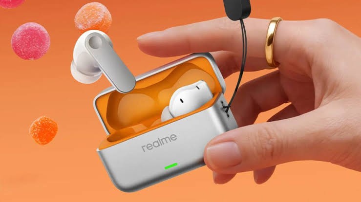 Mão feminina segura um estojo de fones de ouvido Realme cinza e laranja, com um fone branco fora e outro dentro. Um anel dourado está no dedo anelar, e balas de goma coloridas flutuam ao fundo laranja