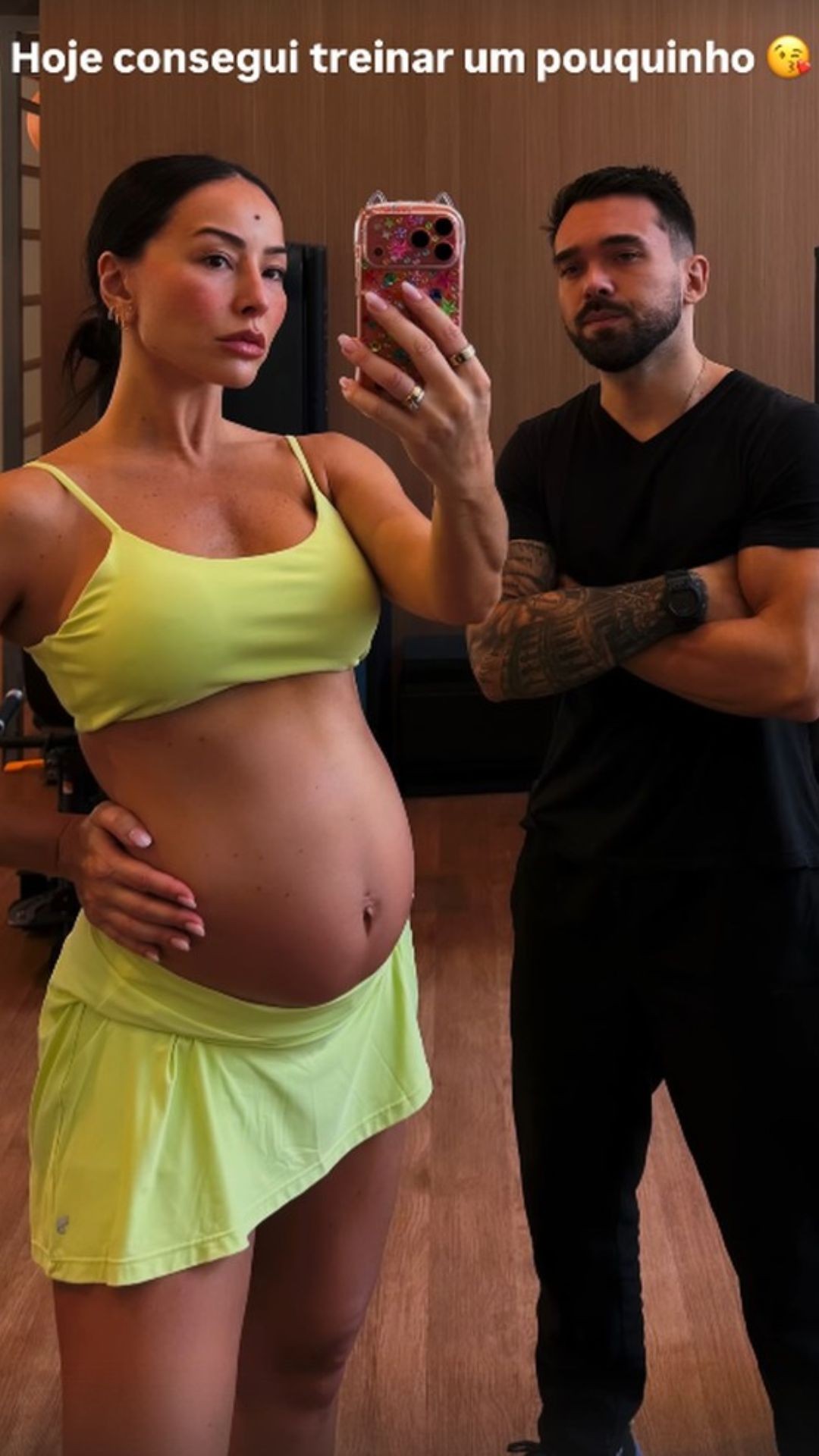 Mulher grávida de perfil, com top e saia verde-limão, segura a barriga enquanto tira uma selfie no espelho. Ao lado, um homem barbudo de camiseta preta e braços cruzados, com tatuagens nos antebraços. O texto na imagem diz: Hoje consegui treinar um pouquinho