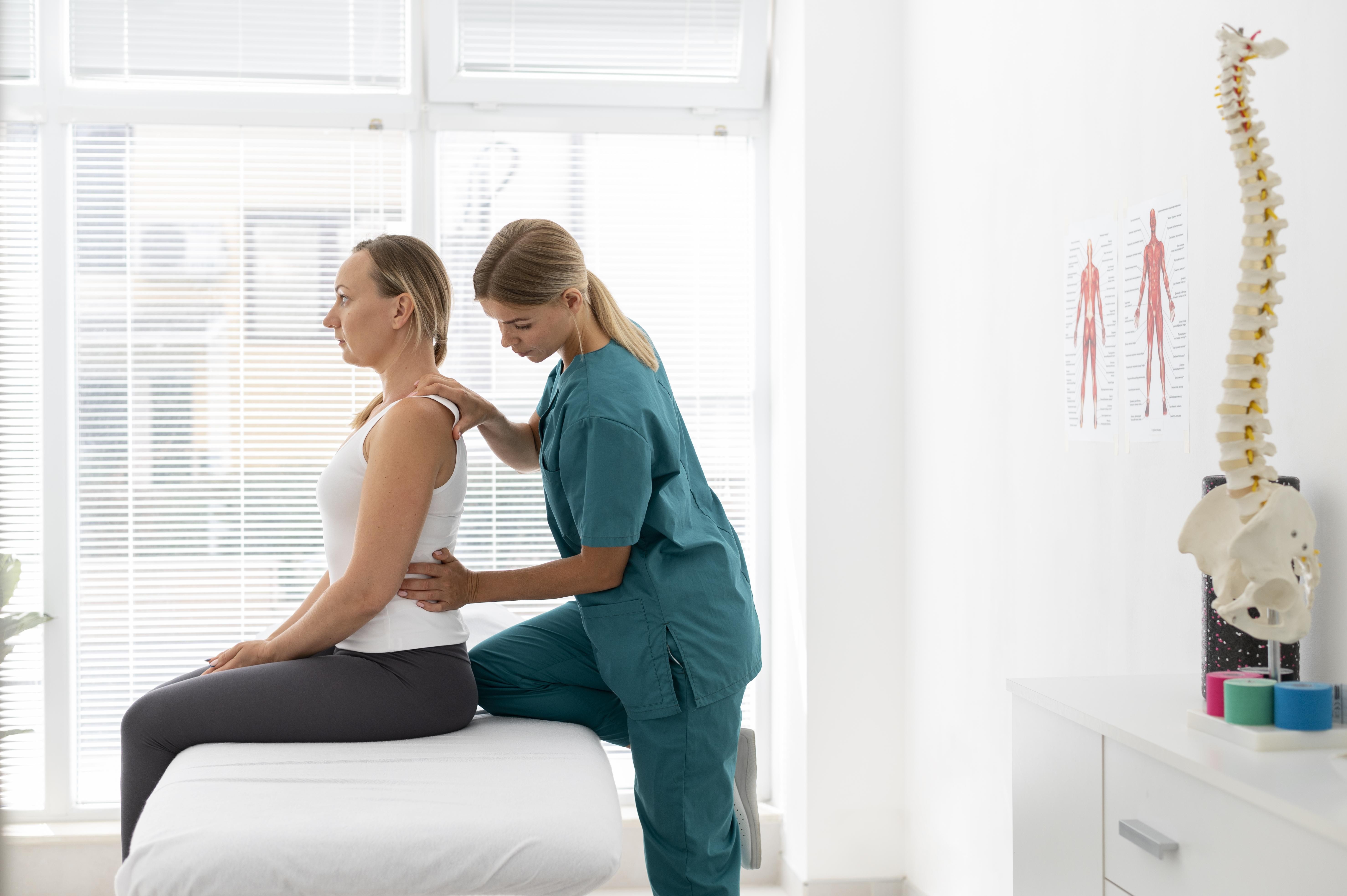 Fisioterapeuta de uniforme verde-azulado examinando a postura de uma mulher sentada em maca branca, com as mãos nas costas e ombro dela. Ao fundo, janela com persianas e um modelo de coluna vertebral em mesa branca