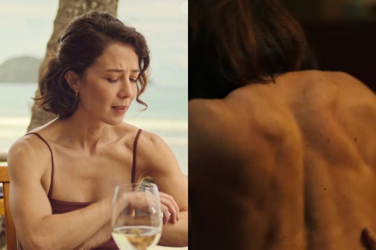 Mulher de cabelo curto e escuro, com expressão séria, usando blusa de alças finas cor vinho, sentada à mesa com taça de vinho branco. À direita, costas musculosas de uma pessoa, com cabelo escuro sobre os ombros, em ambiente de pouca luz