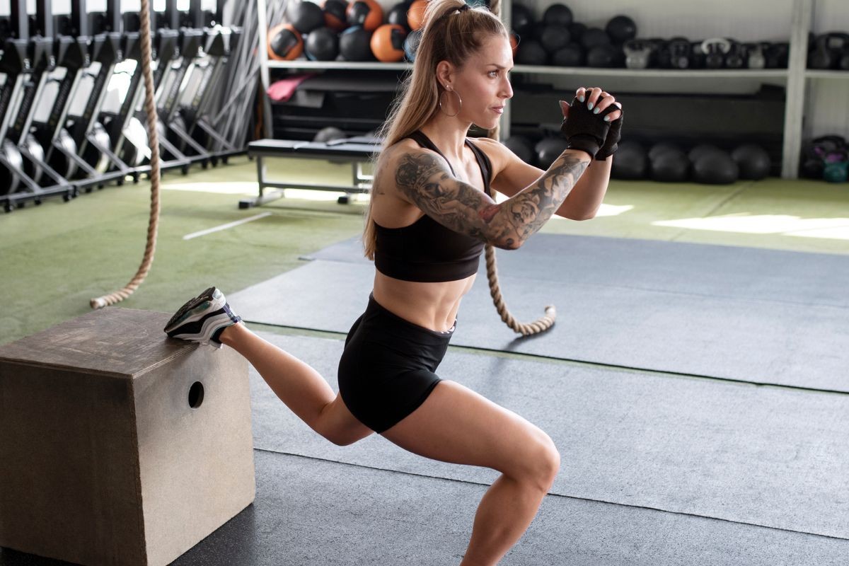 Mulher loira, com tatuagens nos braços, fazendo agachamento búlgaro com o pé esquerdo apoiado em um caixote de madeira em uma academia. Ela veste top e shorts pretos, luvas de treino e tênis esportivos.