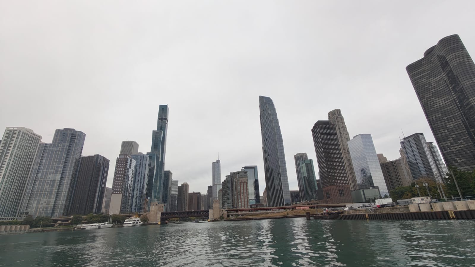 Horizonte urbano de Chicago com arranha-céus modernos e clássicos às margens de um rio, sob céu nublado. Edifícios altos de vidro e concreto dominam a paisagem, com pontes e barcos visíveis na água.