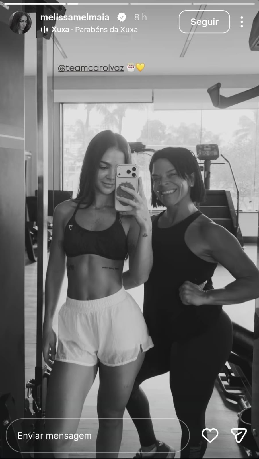 Duas mulheres sorridentes, uma jovem e outra mais velha, posam para uma selfie no espelho de uma academia. A jovem, à esquerda, veste top e shorts brancos, segurando o celular. A mulher mais velha, à direita, veste regata preta e flexiona o braço, exibindo o bíceps. Ambas têm cabelos escuros e curtos, e o fundo mostra equipamentos de ginástica e uma janela com vista para árvores