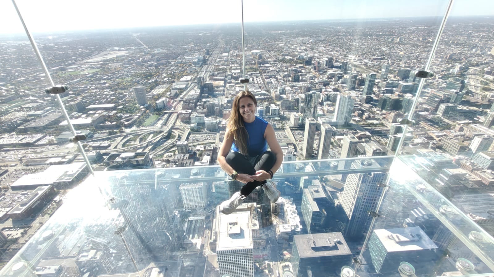 Mulher sorridente de cabelo castanho, blusa azul e calça preta, sentada de pernas cruzadas em uma plataforma de vidro transparente, com a cidade vista de cima, repleta de edifícios e ruas, sob um céu claro