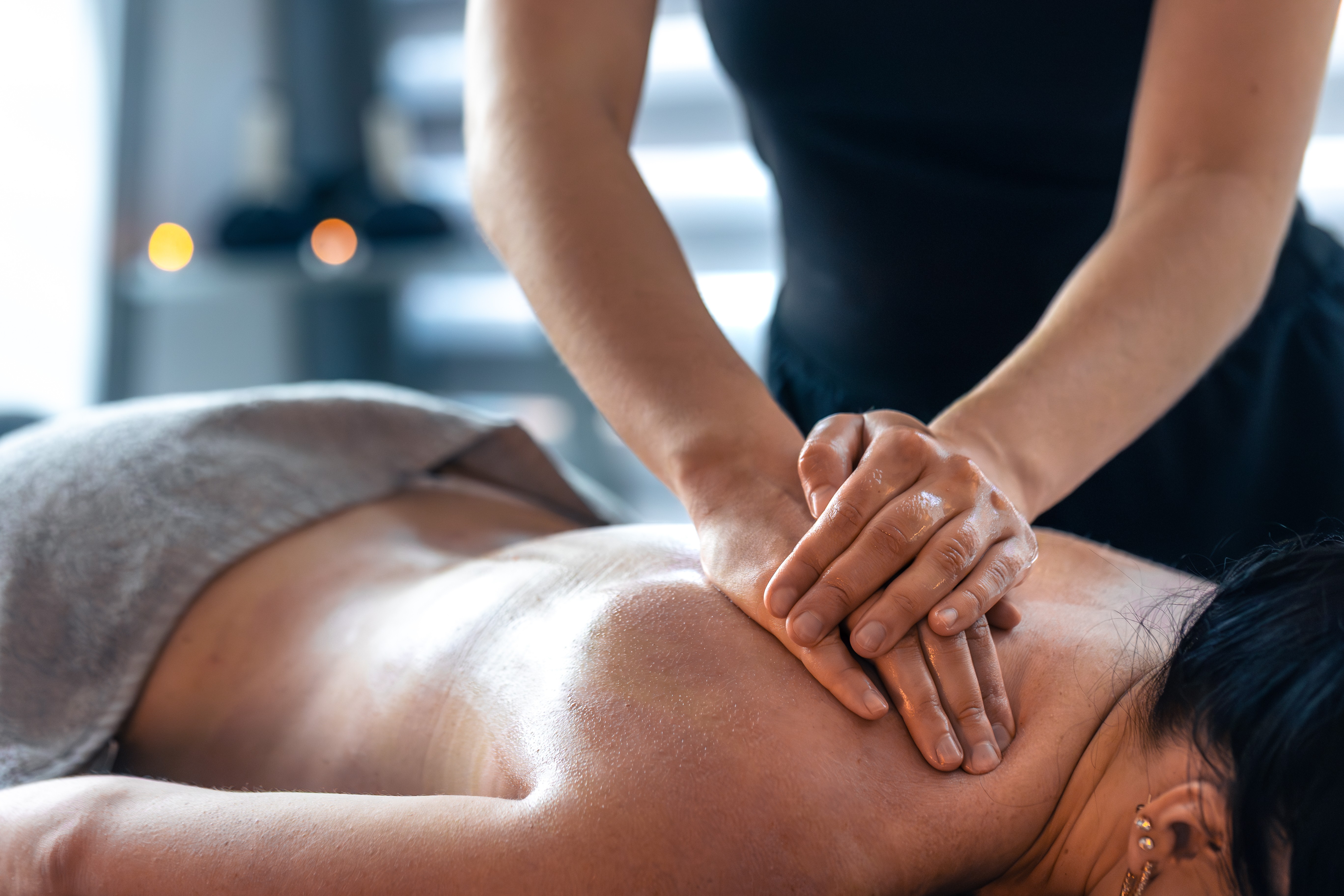Uma pessoa deitada de bruços, com as costas nuas e brilhantes de óleo, recebendo uma massagem relaxante. As mãos do massagista, também com óleo, estão sobre o ombro esquerdo da pessoa, aplicando pressão. Um tecido cinza cobre a parte inferior do corpo. O ambiente é suavemente iluminado, com luzes desfocadas ao fundo