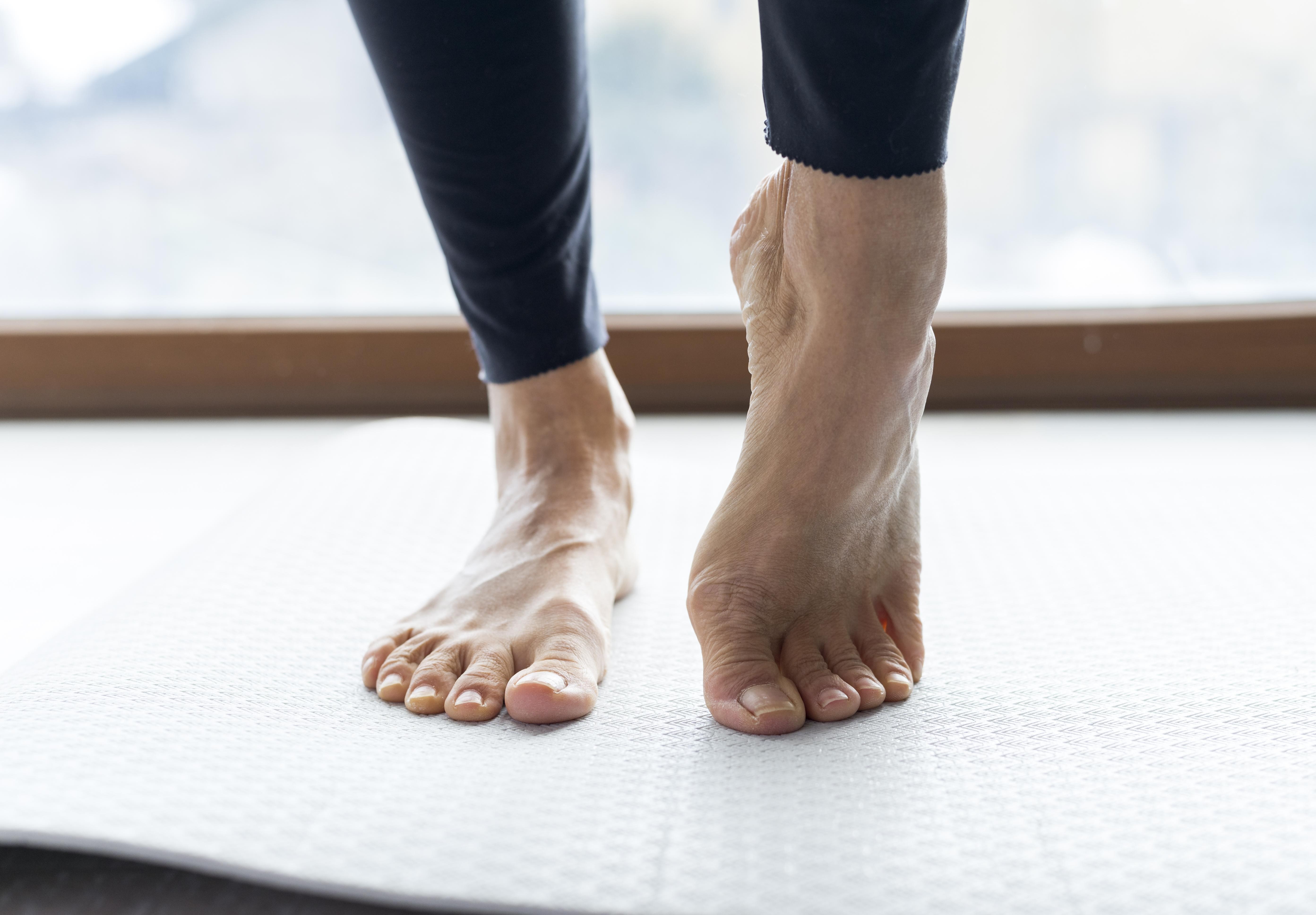 Pés de uma pessoa em leggings pretas sobre um tapete de yoga cinza, com o pé direito na ponta e o esquerdo totalmente apoiado, em frente a uma janela com luz natural